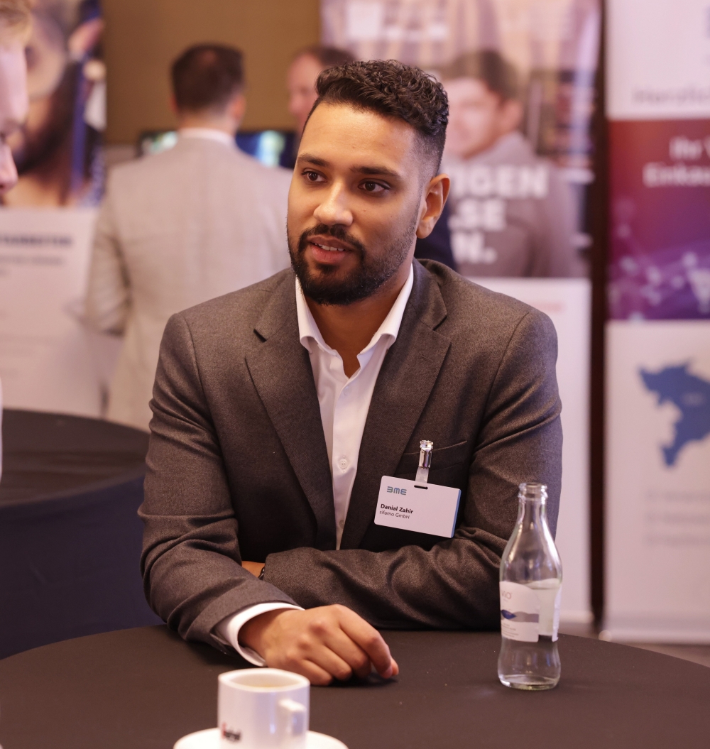 Mann mit dunklem Bart und lockigem Haar in grauem Anzug sitzt an einem Tisch mit Kaffeetasse und Wasserflasche.