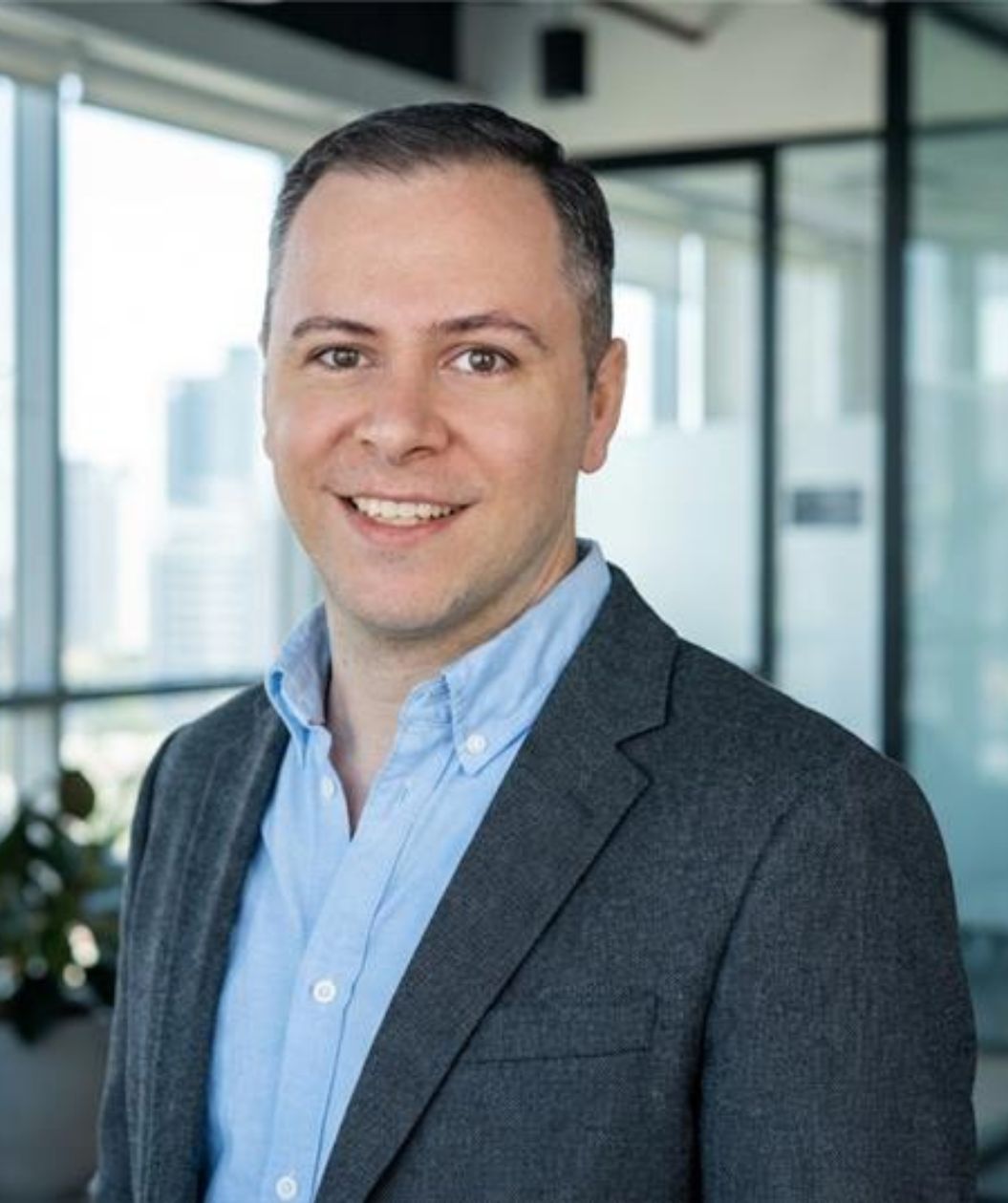 Lächelnder Mann in grauem Blazer und hellblauem Hemd vor großen Fenstern in einem modernen Büro.