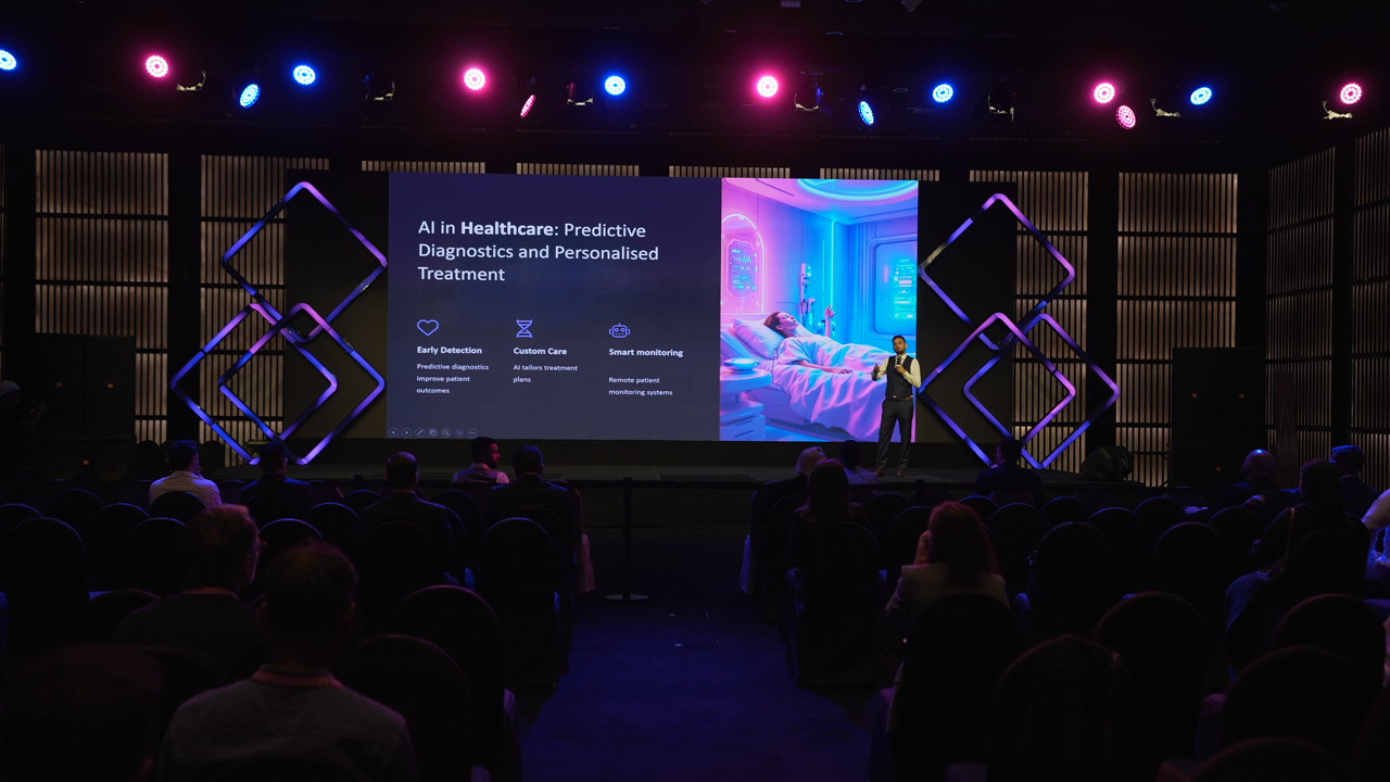 Ein Redner präsentiert eine Folie über KI im Gesundheitswesen auf einer dunklen Bühne vor Publikum.