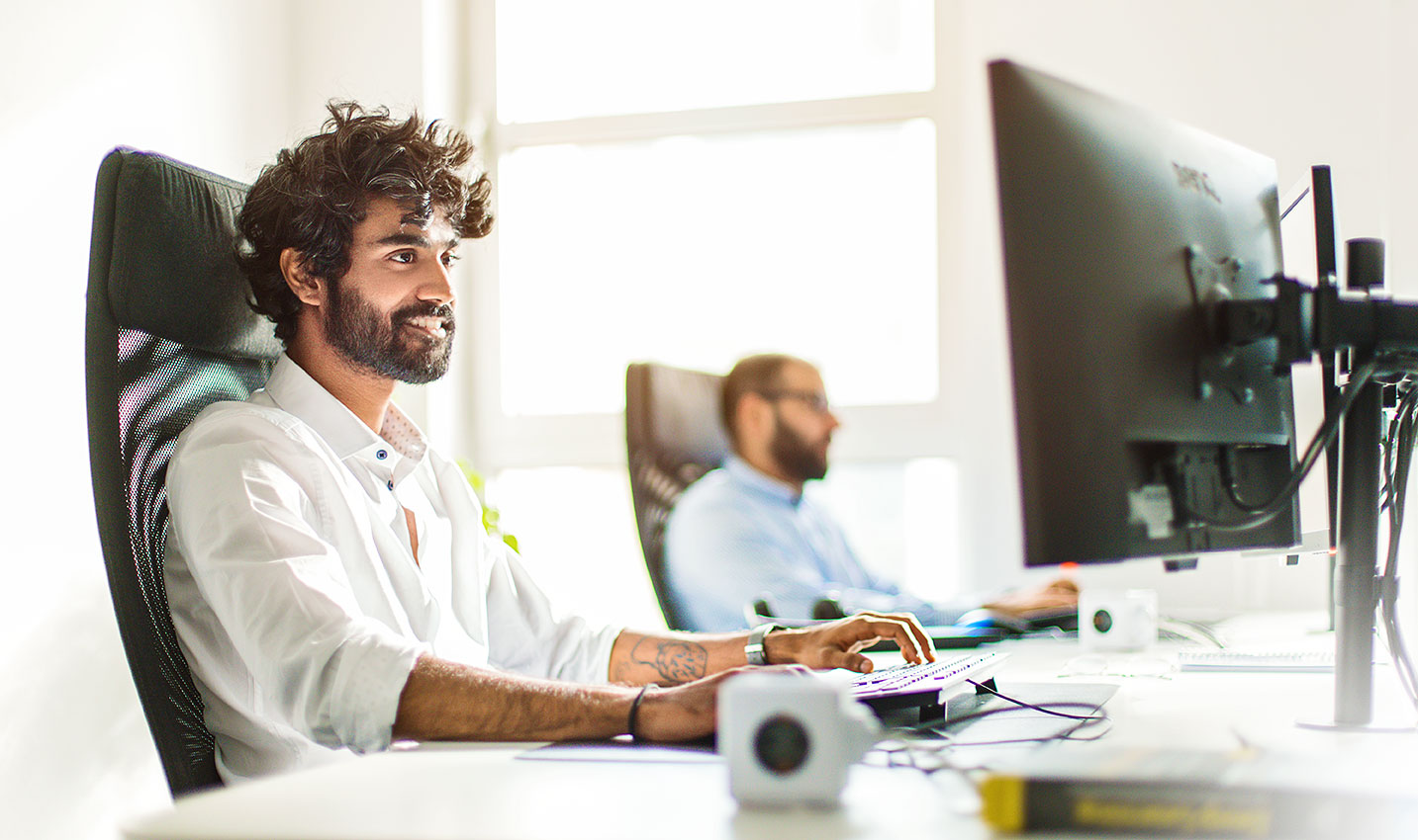 Zwei Männer arbeiten an Schreibtischen mit Computern in einem hellen Büro.