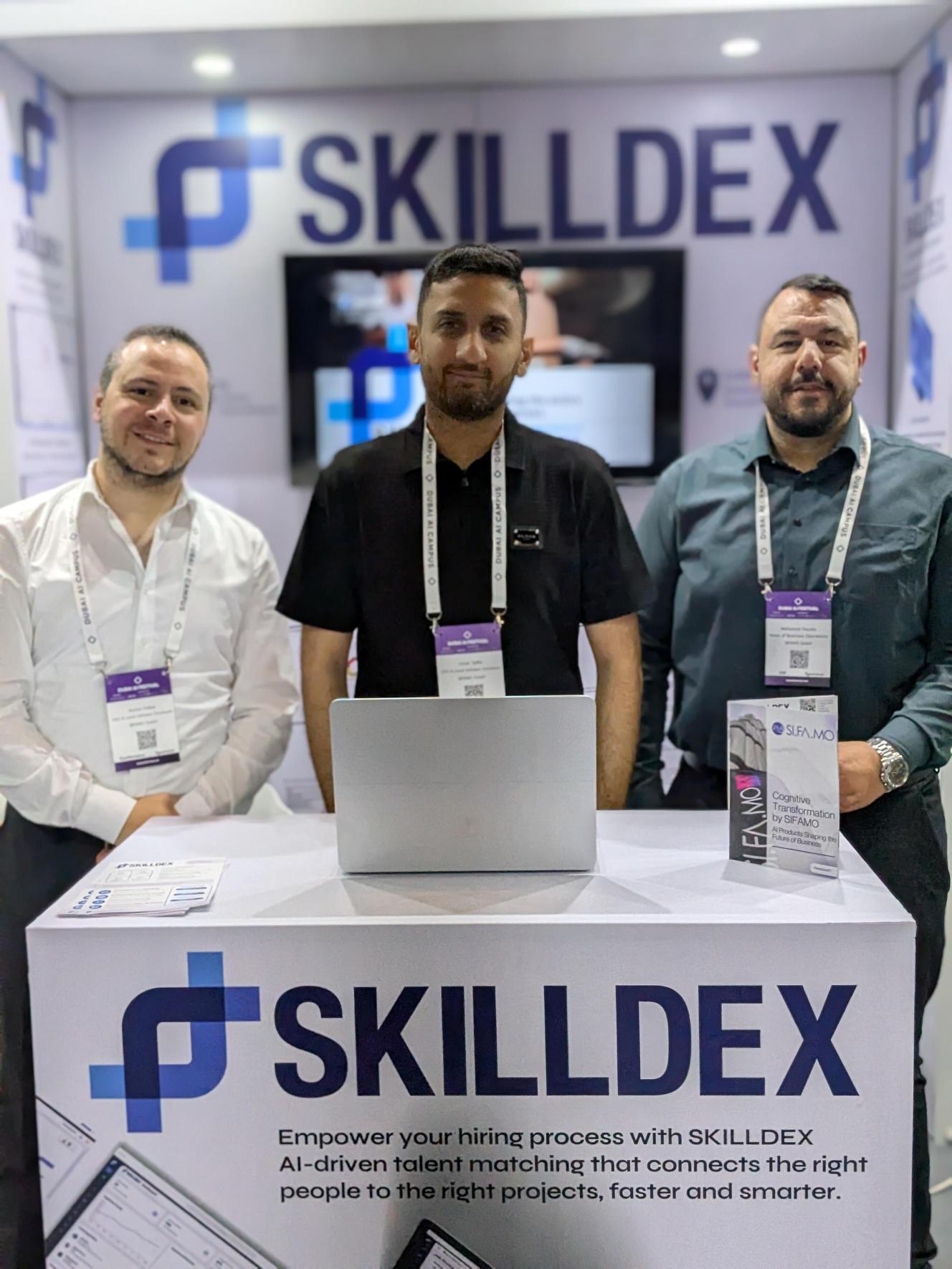 Drei Personen stehen hinter einem Messestand mit dem Schriftzug SKILLDEX und einem Laptop auf dem Tisch.